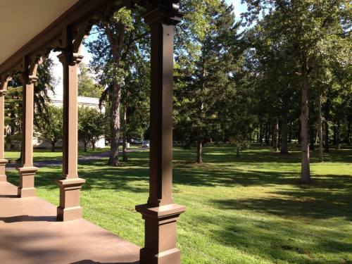 A view from a porch with wooden columns, overlooking a grassy area and trees in a sunny park setting.