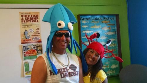 A man in a blue squid hat and a woman in a red crab hat smile in a colorful room with fish posters.