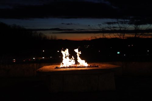 A fire burns brightly in a stone pit against a darkening sky at sunset, with distant lights twinkling in the background.