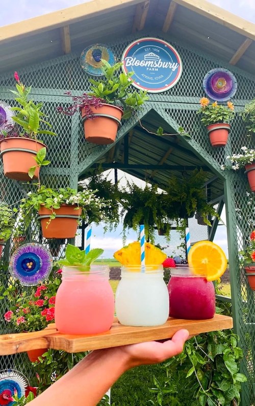 A hand holds a wooden tray with four colorful drinks in front of a floral-decorated gazebo.