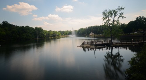 A serene lake scene with a fountain, surrounded by lush trees and a wooden dock under a partly cloudy sky.