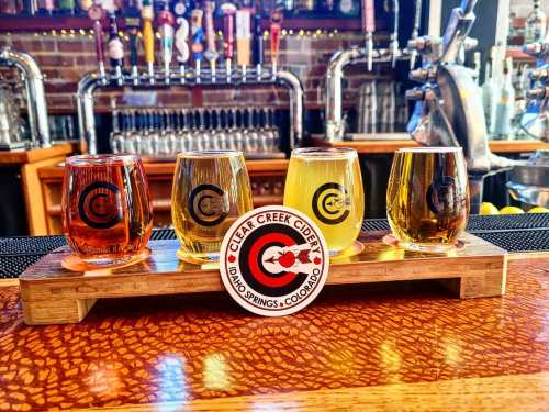 A wooden flight board with four glasses of cider in different colors, featuring a logo for Clear Creek Cidery.