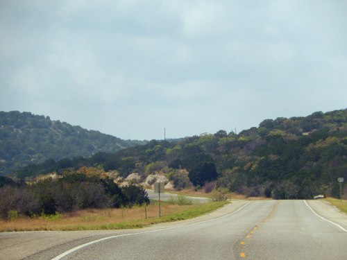 A winding road stretches through green hills under a cloudy sky, with trees lining the sides.