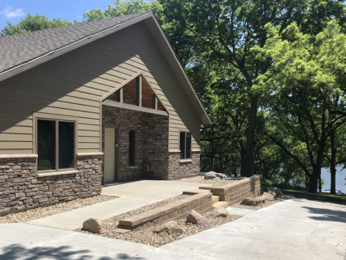 A modern house with a stone facade, surrounded by trees, near a lake on a sunny day.