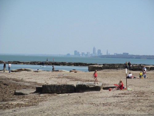 A sandy beach with people enjoying the sun, rocky formations, and a distant city skyline under a clear blue sky.