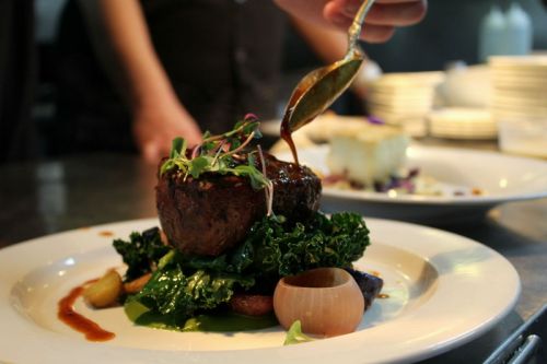 A gourmet dish featuring a beef filet topped with sauce, served on a bed of kale and garnished with microgreens.