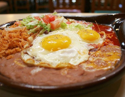 A plate of Mexican food featuring rice, refried beans, two sunny-side-up eggs, and fresh salad toppings.