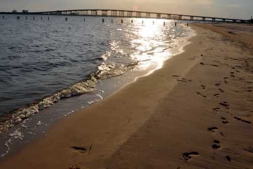 A sandy beach with footprints, gentle waves, and a bridge in the distance under a glowing sunset.