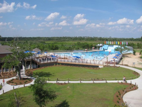 Aerial view of a large swimming pool area with people swimming, surrounded by green grass and colorful flags in the background.