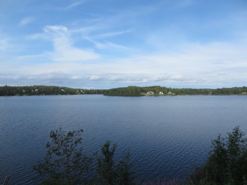 A serene lake surrounded by lush greenery and distant houses under a blue sky with scattered clouds.