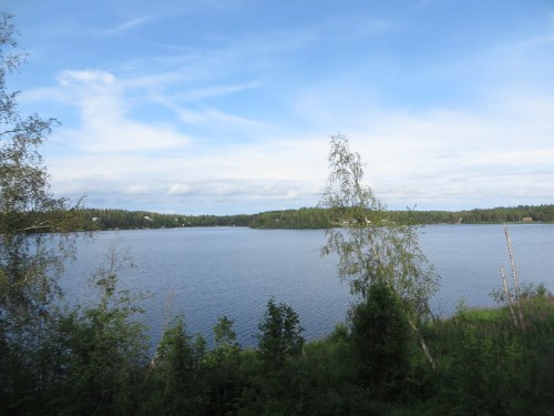 A serene lake surrounded by lush greenery and trees under a blue sky with scattered clouds.
