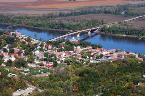 Aerial view of a small town by a river, featuring colorful houses, a bridge, and lush greenery in the surrounding landscape.