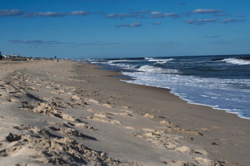 A sandy beach with gentle waves, under a clear blue sky dotted with clouds.