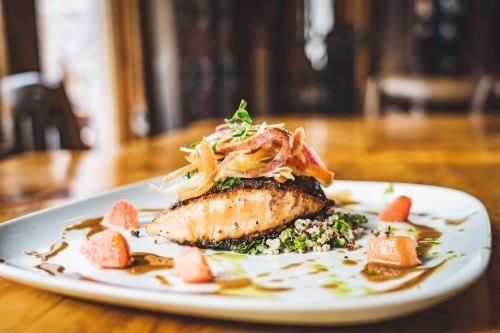 A beautifully plated salmon dish topped with greens, served on a bed of quinoa and garnished with fruit and sauce.