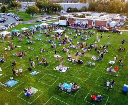 Aerial view of a large outdoor gathering with people sitting on blankets and chairs, surrounded by tents on a grassy field.