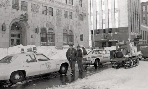 Two men stand in front of a police station with snow-covered streets and parked cars, including a police vehicle.