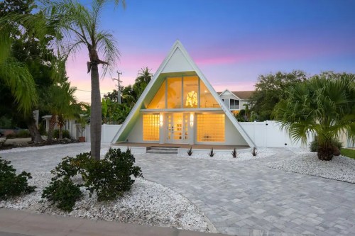 A modern A-frame house with large windows, surrounded by palm trees and a landscaped yard at sunset.