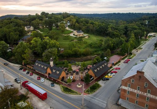 Aerial view of a scenic town with green hills, vineyards, and historic buildings along a road.