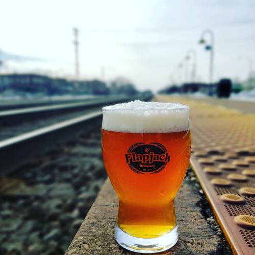 A glass of amber beer with a frothy head sits on a platform near train tracks, featuring the Flapjack Brewery logo.