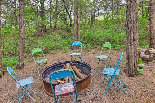 A fire pit surrounded by colorful, weathered chairs in a wooded area with lush greenery.
