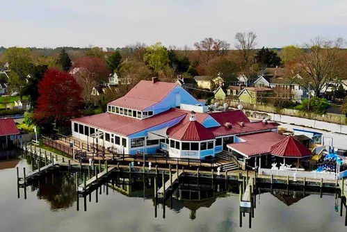 Aerial view of a waterfront restaurant with a red roof, surrounded by trees and houses, reflecting in the water.