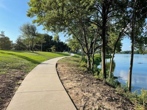A winding concrete path beside a calm lake, surrounded by green grass and trees under a clear blue sky.
