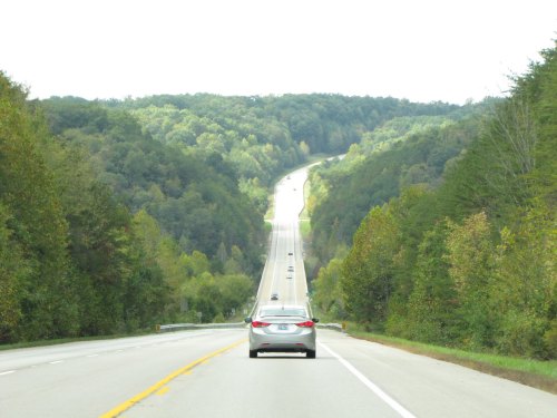 A car drives down a winding highway surrounded by lush green hills under a cloudy sky.
