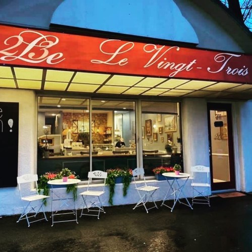 A cozy café exterior with a red sign reading "Le Vingt-Trois," featuring outdoor seating and flower pots.
