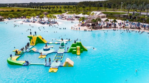 A vibrant water park scene with a large inflatable play structure in a blue lagoon, surrounded by people enjoying the sun.