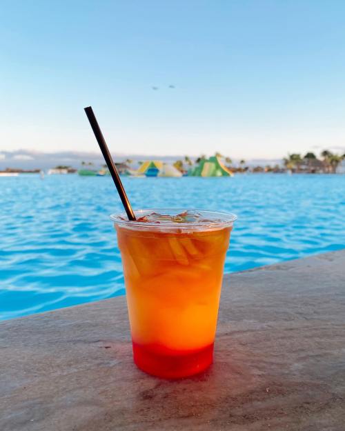 A colorful drink with ice sits on a ledge by a bright blue pool, with inflatable structures in the background.