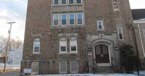 Brick building with multiple windows and a prominent entrance, set against a cloudy sky and snow-covered ground.