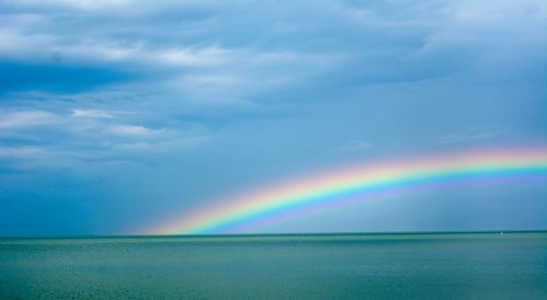A vibrant rainbow arcs over a calm sea under a cloudy blue sky.