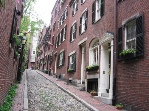 A narrow, cobblestone street lined with historic brick buildings and flower boxes in a charming neighborhood.