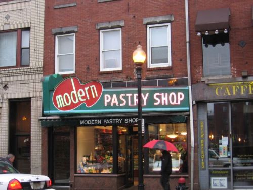 A storefront of "Modern Pastry Shop" with a bright sign, surrounded by brick buildings and a lamppost.