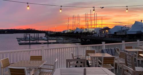 A serene waterfront view at sunset, featuring boats, a marina, and string lights illuminating outdoor tables.