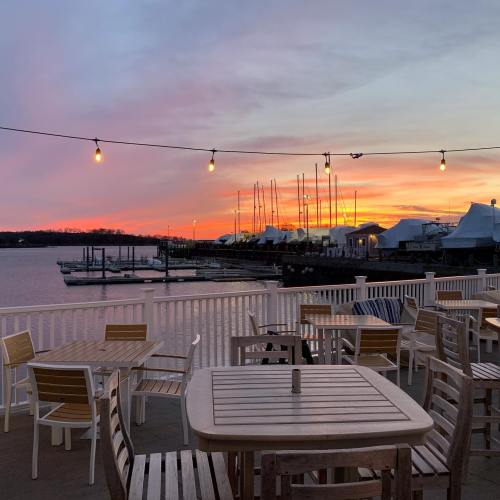 A serene waterfront view at sunset, featuring boats, a marina, and string lights illuminating empty tables.
