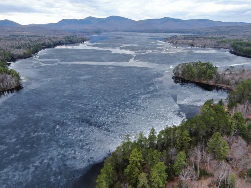 Aerial view of a frozen lake surrounded by trees and mountains under a cloudy sky.