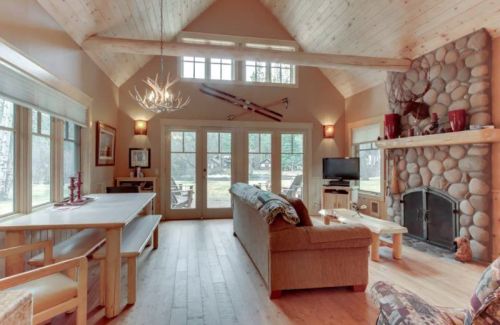 Cozy living room with high ceilings, wooden beams, stone fireplace, and large windows overlooking a natural landscape.