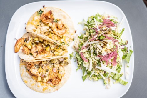 Three shrimp tacos topped with corn and herbs, served with a side salad of lettuce, cabbage, and pumpkin seeds.