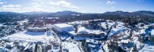 Aerial view of a snowy landscape featuring buildings, mountains, and a clear blue sky.