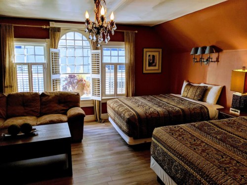 Cozy hotel room with two beds, a sofa, a chandelier, and large windows letting in natural light. Warm, inviting decor.
