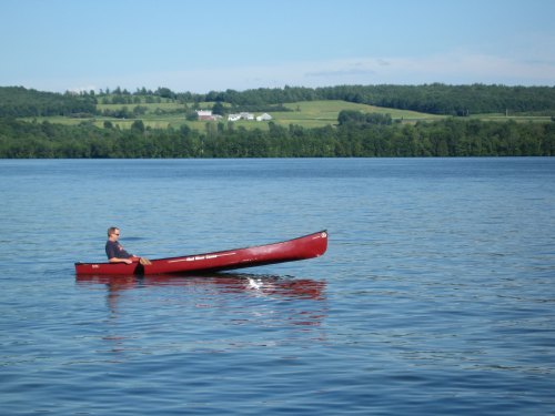 A person paddles a red canoe on a calm lake, surrounded by green hills and a clear blue sky.