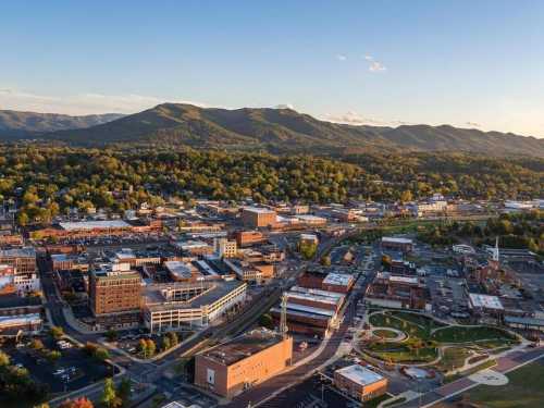 Aerial view of a vibrant town surrounded by green mountains, showcasing buildings, parks, and a clear blue sky.