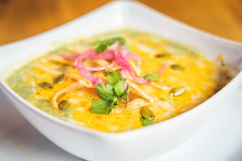 A bowl of creamy soup topped with cheese, crispy tortilla strips, pickled onions, and fresh cilantro.