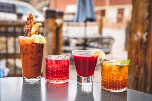 Four colorful cocktails on a table: a Bloody Mary, a pink drink, a margarita, and an orange cocktail, set outdoors.