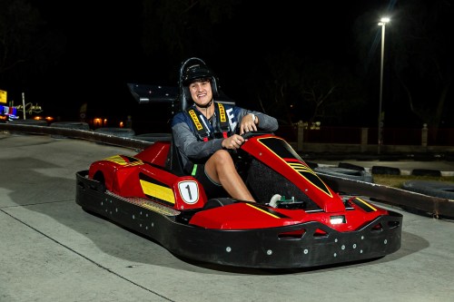 A driver in a go-kart smiles while sitting on a racetrack at night, wearing a helmet and racing gear.