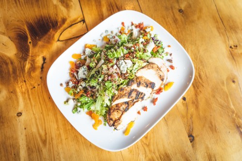 Grilled chicken salad with mixed greens, cheese, and colorful vegetables on a white plate, set on a wooden table.