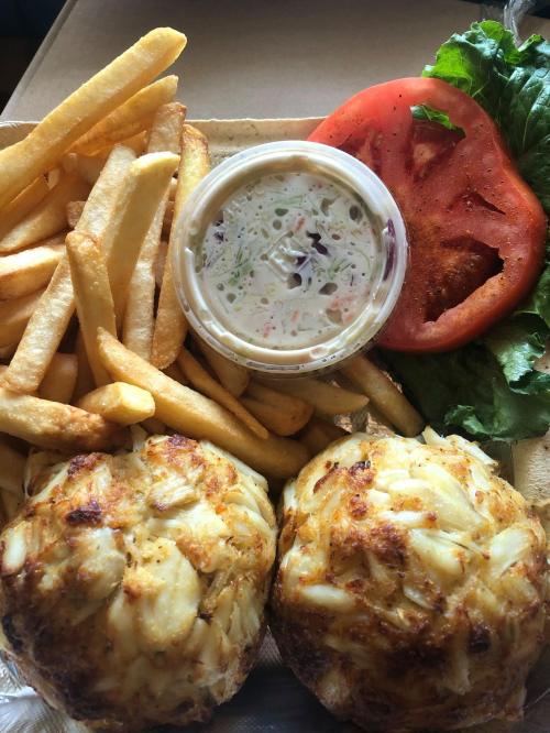 A takeout box with two crab cakes, fries, a slice of tomato, lettuce, and a small container of dipping sauce.