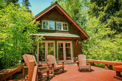 A cozy cabin surrounded by trees, featuring a wooden deck with Adirondack chairs and large windows.