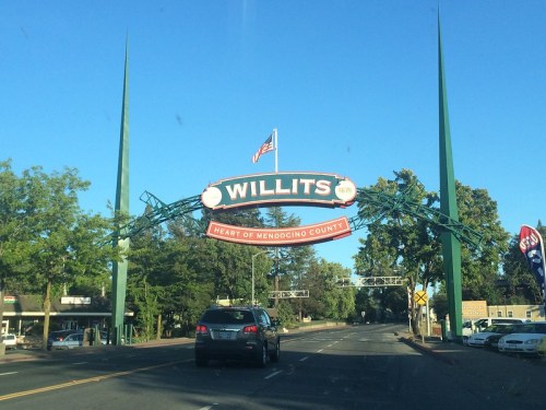 Sign welcoming visitors to Willits, California, with green spires and an American flag, set against a clear blue sky.